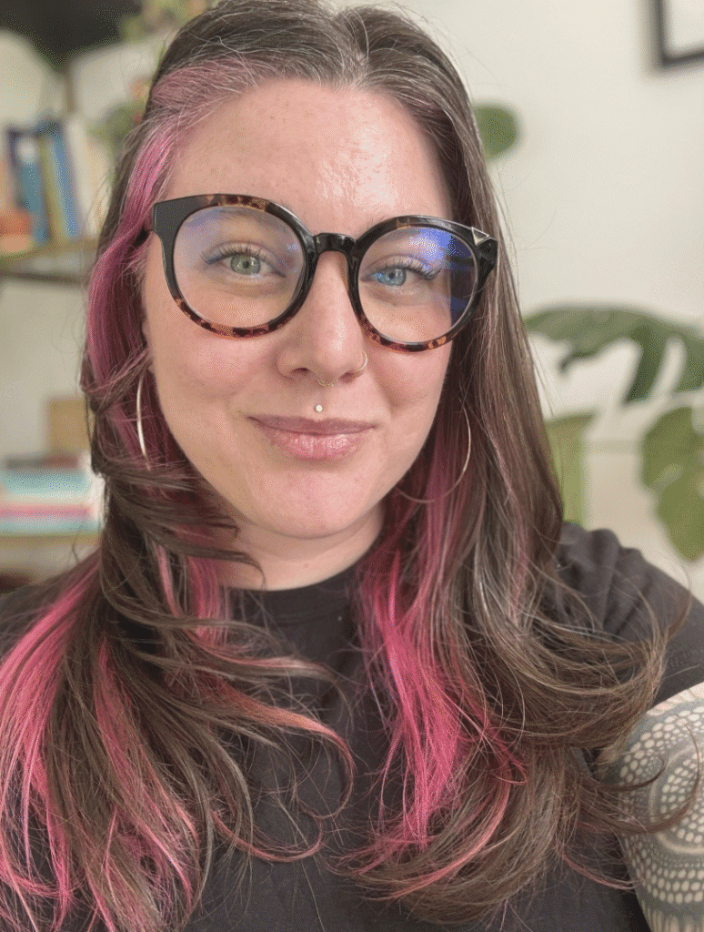 Photo portrait of a fair skinned person smiling. She has tortoise shell glasses, and brown hair with a bright pink streak